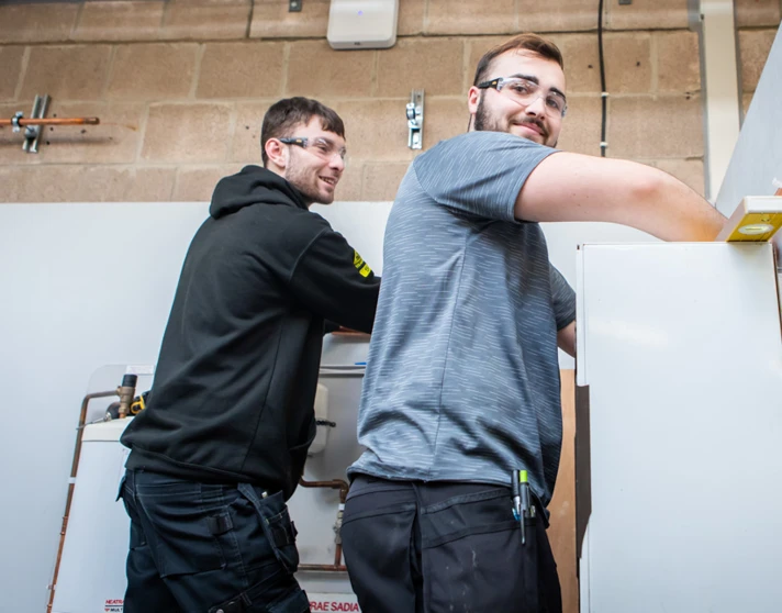 Two plumbing students smiling while collaboratively working on installing a plumbing fixture, engaged in hands-on training. Two plumbing students smiling while collaboratively working on installing a plumbing fixture, engaged in hands-on training.